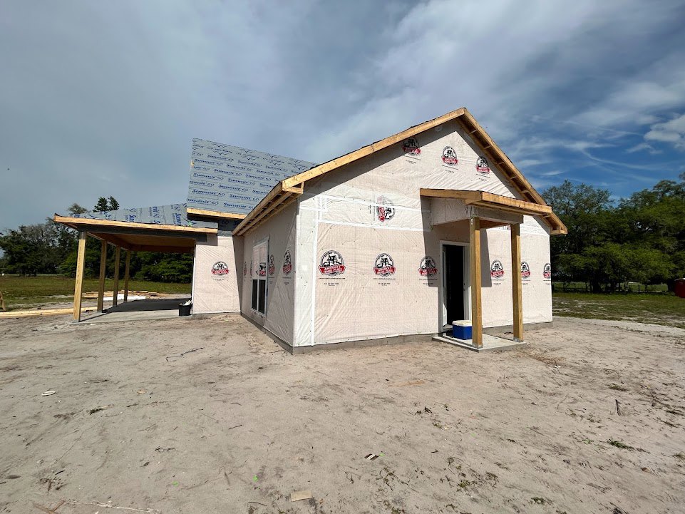 White custom home under construction with completed wooden roof, exposed framing, sandy ground, and surrounding trees under cloudy sky
