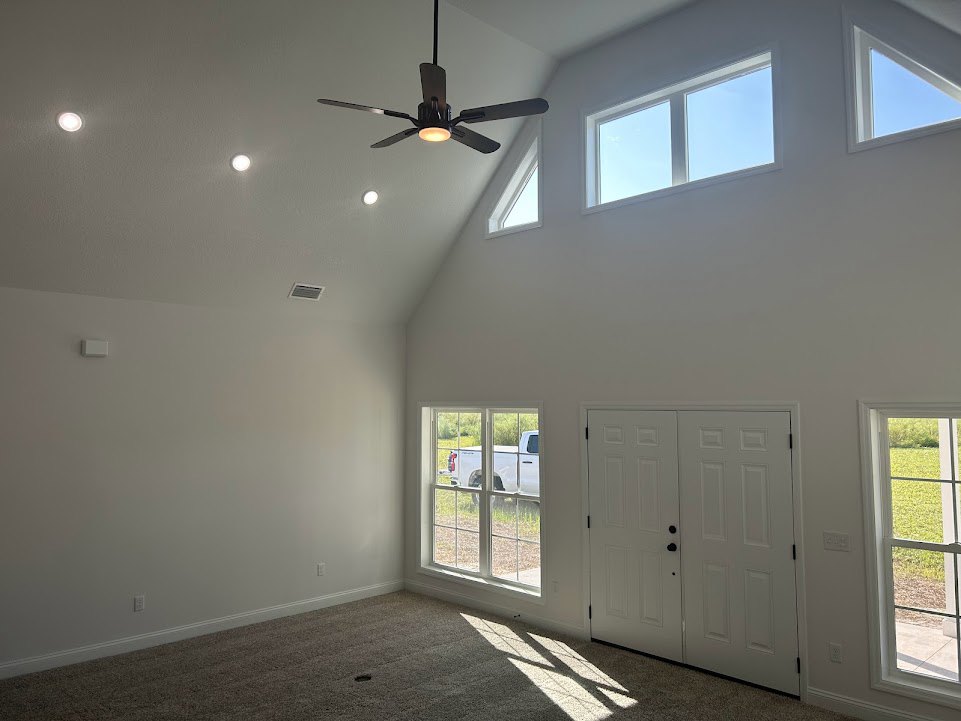 Ceiling fan with integrated light fixture mounted on white ceiling, flanked by double doors with black knobs and windows showing blue sky and grassy field.