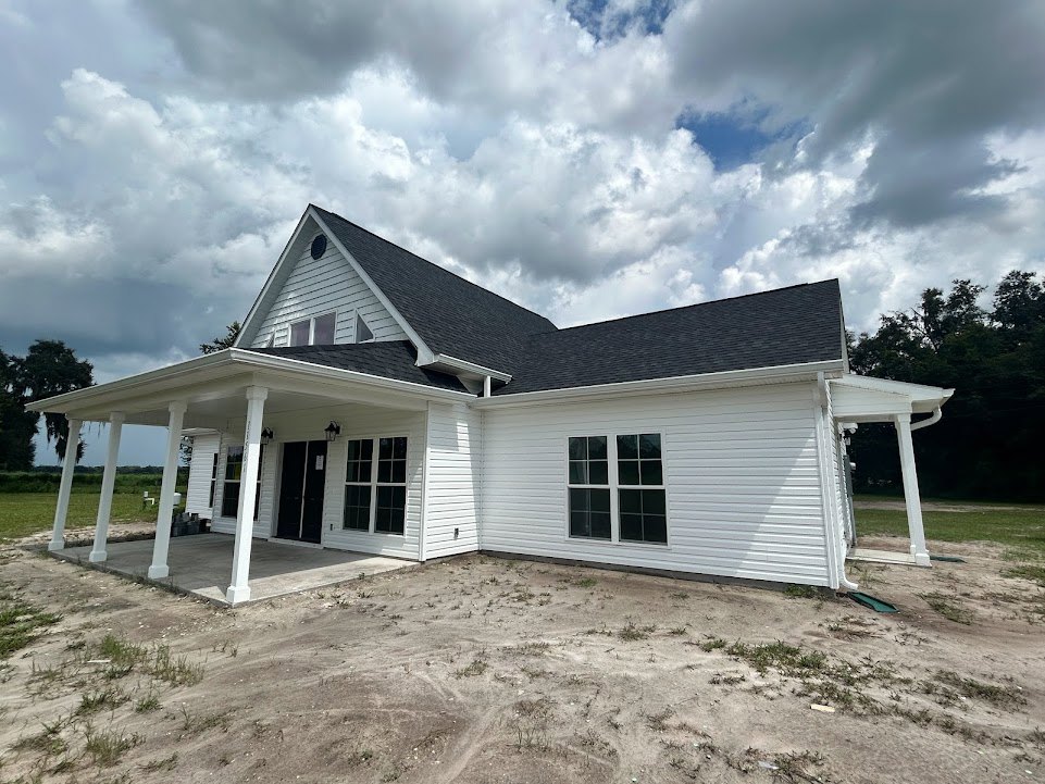 Two-story house with black roof, white siding, multiple windows, black front door, covered porch, large grassy yard with dirt patch, trees, and partly cloudy sky