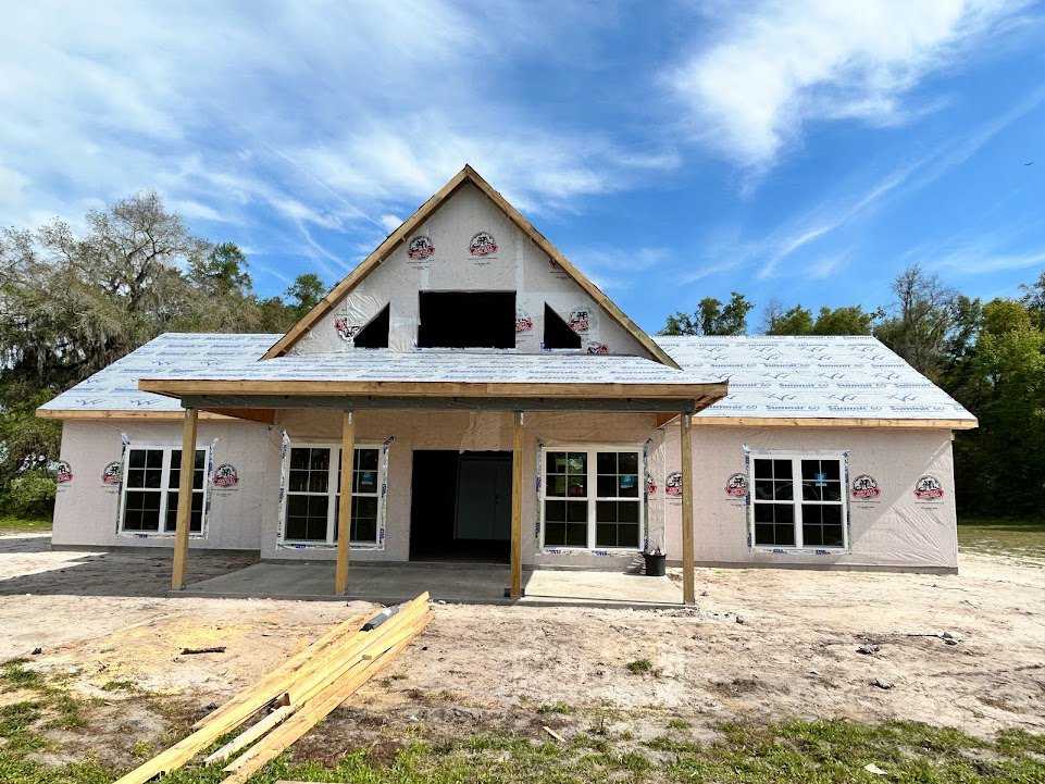 Partially built house with shingled roof, multi-pane windows, unfinished doorway, and stacked lumber on dirt lot, surrounded by trees under cloudy sky
