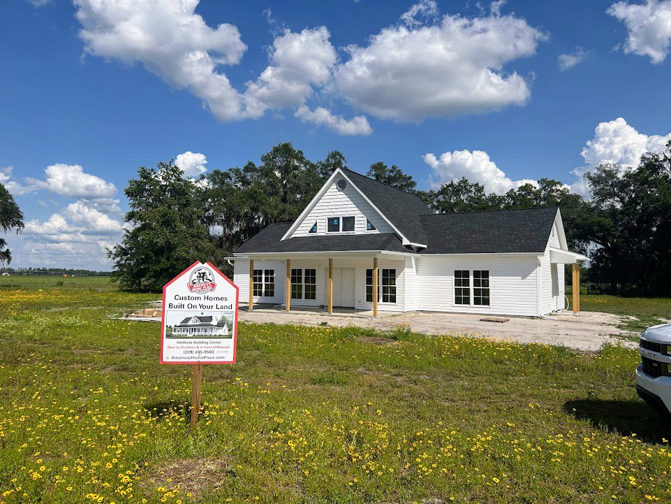 Two-story house with black roof, white siding, and large front windows, manicured lawn, blue sky with scattered clouds, real estate sign near driveway, parked car partially visible