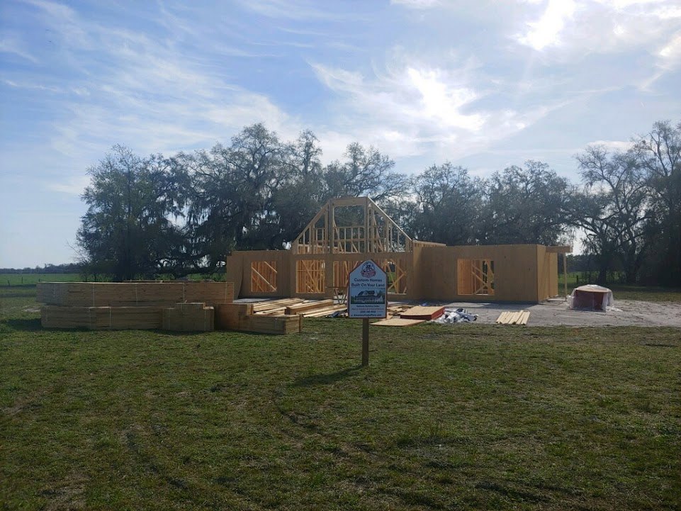 Wood-framed house under construction with exposed beams and staircase, grassy lot, construction sign featuring a house illustration, scattered trees and cloudy sky in background