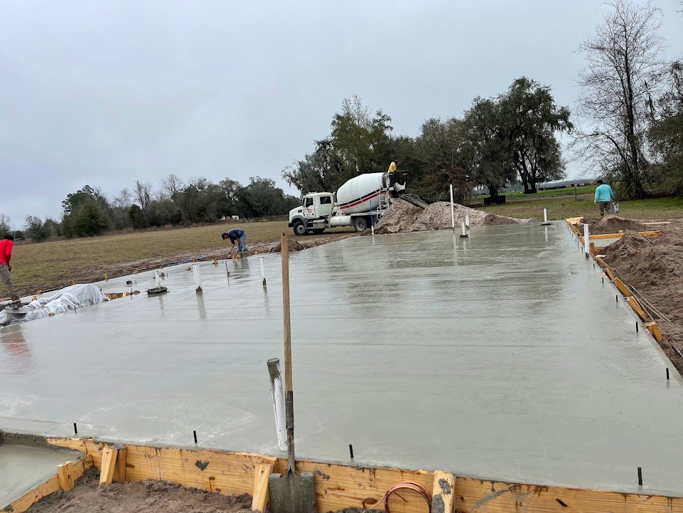 Freshly poured concrete slab with a cement mixer truck and worker, bare tree in background, metal pole in foreground, outdoor construction site under clear sky