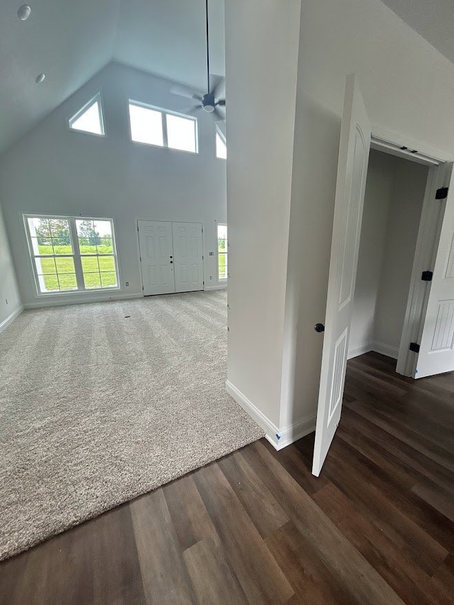 Bedroom with beige carpet, white double doors with black knobs, ceiling fan, window overlooking grassy field, wood flooring in adjacent area, wall-mounted light fixture