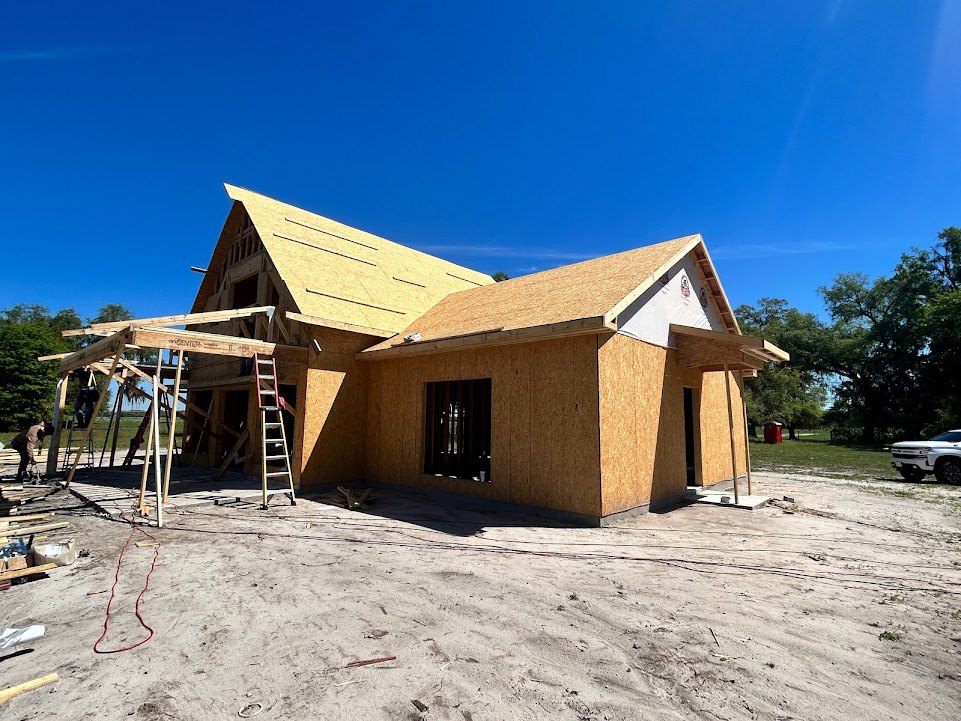 Wood-framed house under construction with exposed beams, ladder leaning against exterior, white truck parked nearby, sunlight illuminating window, red rope on white surface, trees