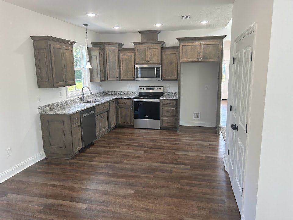 Wood cabinets and matching wood floor in a kitchen with a black and silver oven, built-in microwave, white walls, and white electrical outlet