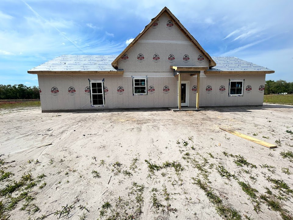 Partially built house with gray shingle roof, white-framed windows, wooden porch, white front door, surrounded by dirt field with patches of grass and scattered lumber