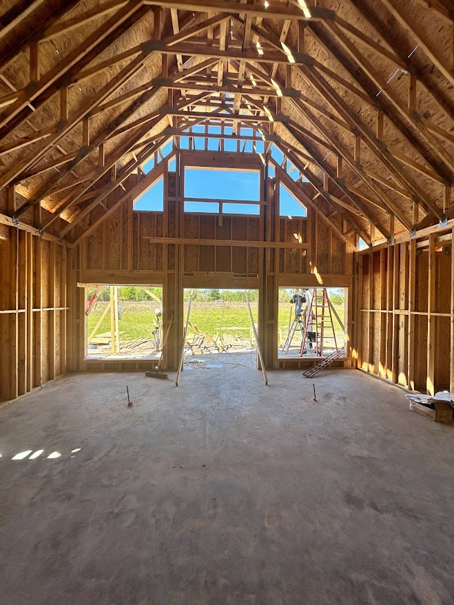 Exposed wooden ceiling beams, concrete floor with embedded metal rods, large window framing a grassy field and blue sky, unfinished walls