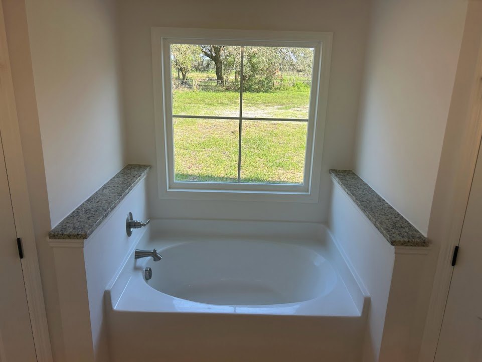 Freestanding white bathtub beneath a large window overlooking green grass and trees, surrounded by light tile walls and modern chrome faucet