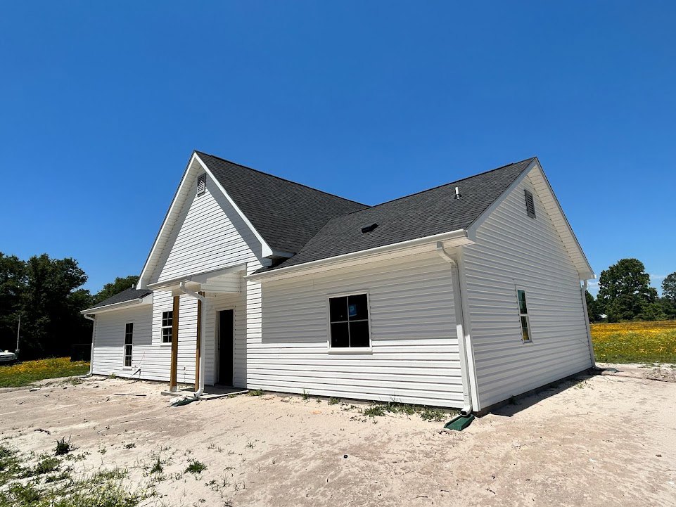 White house with white siding under construction, open front door, window displaying a sign, leafy tree nearby, gray shingled roof visible against blue sky