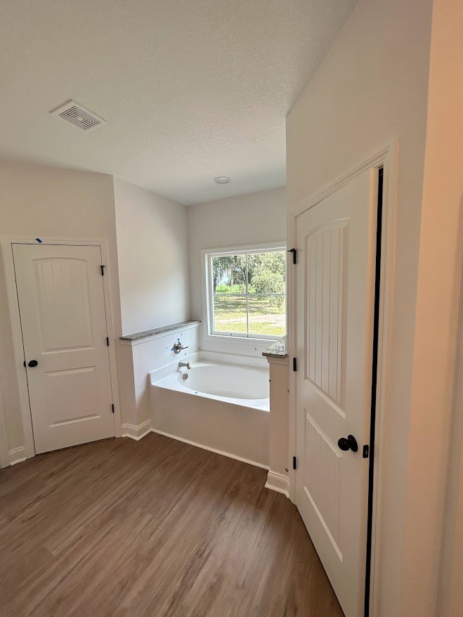 Freestanding white bathtub beneath a large window overlooking a grassy field, wood laminate flooring, white door with black handles, ceiling vent, light-colored walls