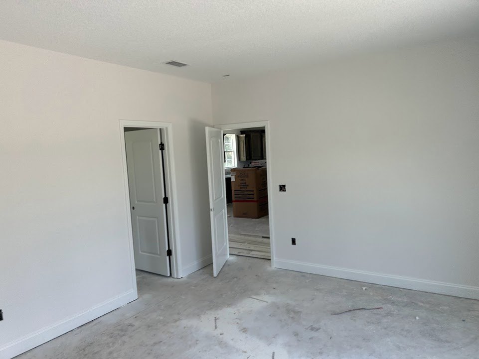 White-walled room with open white door featuring black hinges, white flooring, and a box with a red stripe near the doorway; close-up of wood plank visible.