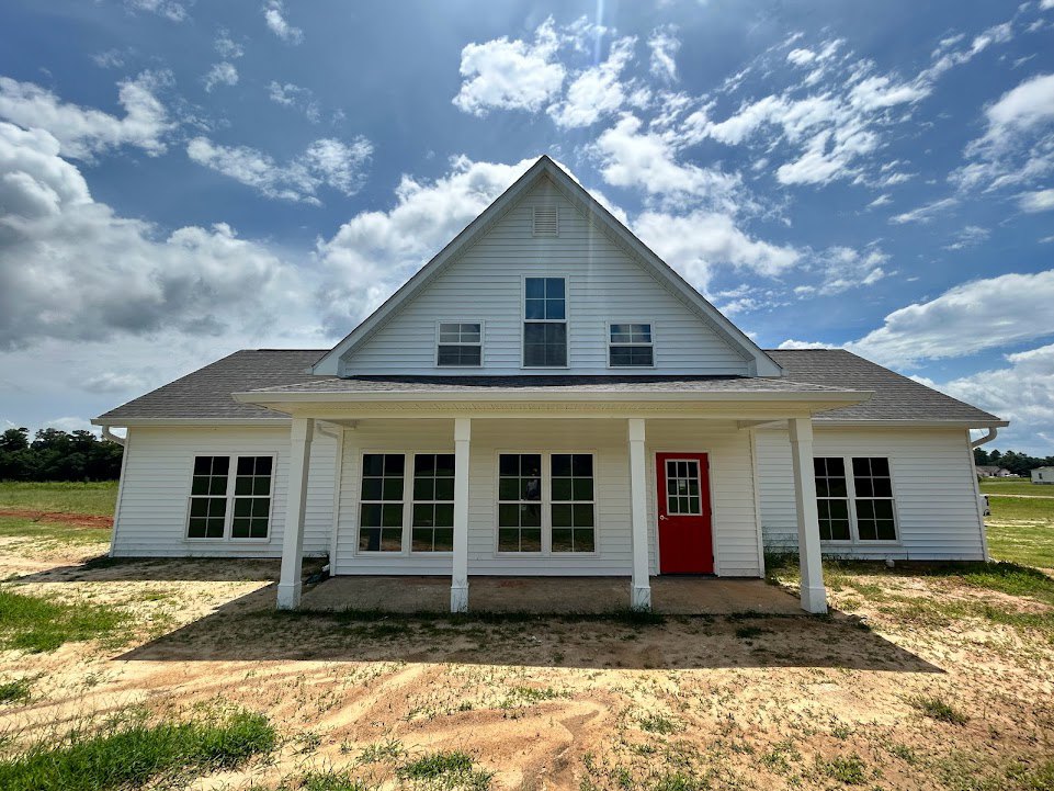 White siding house with triangular roof, red front door with white trim, front porch, grassy lawn, large windows, partly cloudy sky