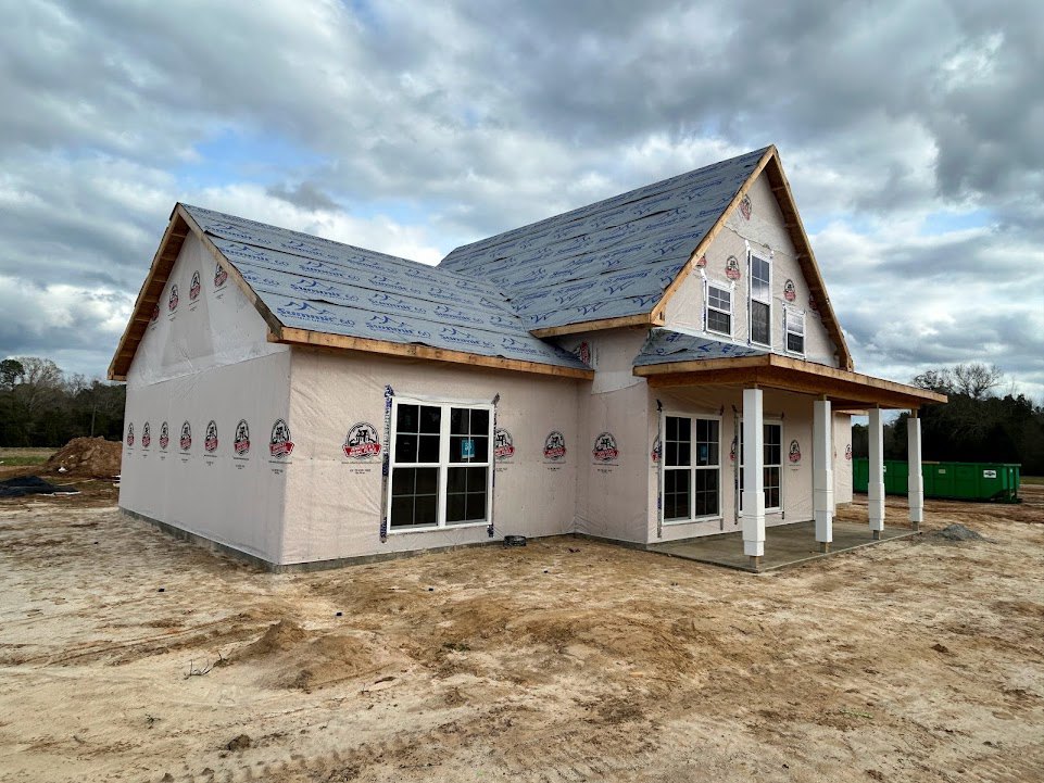Partially built house with exposed wooden framing, unfinished roof, dirt ground, white door displaying a sign, and cloudy sky overhead
