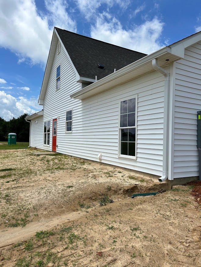 White siding house with red front door, white-framed windows, dirt yard, and green container near trees under cloudy sky
