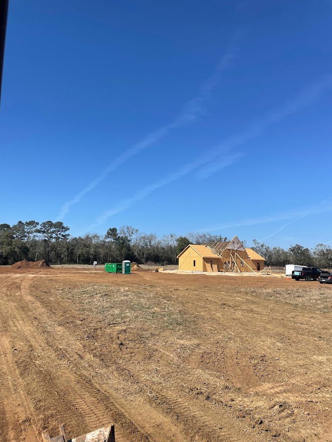 Partially built yellow house with exposed framing and roof, surrounded by dirt field and green dumpster, under blue sky with scattered clouds