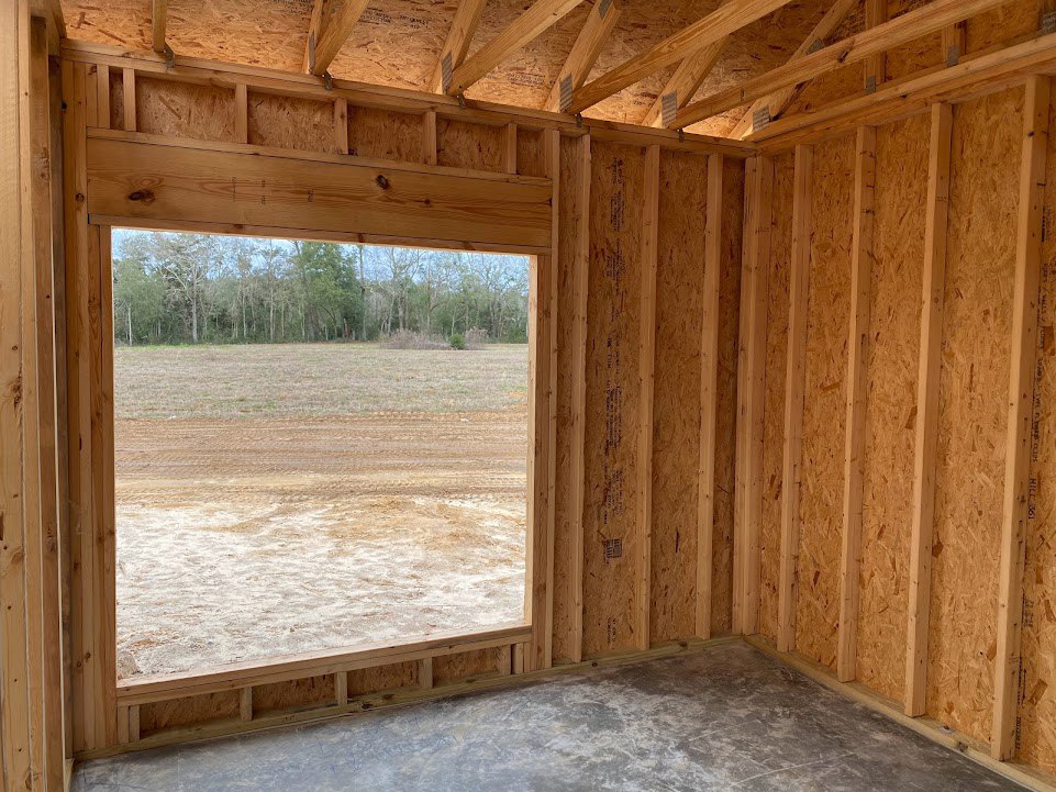 Unfinished room with exposed wood framing and concrete floor, large window overlooking dirt field and trees