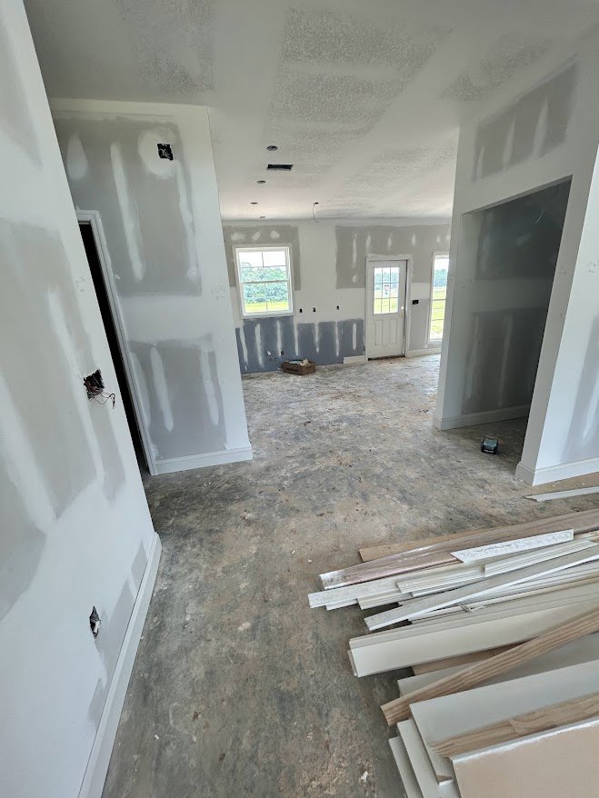 White-walled room with wood plank flooring, white door featuring a window, wall opening, window overlooking forest, stacks of white plastic strips and boards.