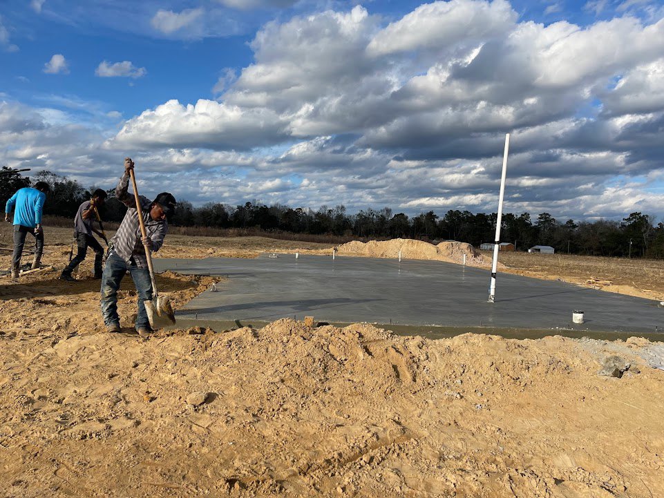 Construction workers in boots and blue shirts digging and shoveling soil on a dirt-filled residential site, tire tracks visible, white pole near a lake, cloudy sky overhead, trees