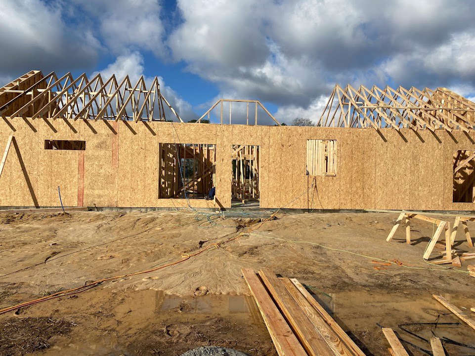 Wood-framed house under construction with exposed beams, pile of lumber on dirt ground, brick exterior partially visible, blue sky with scattered clouds overhead