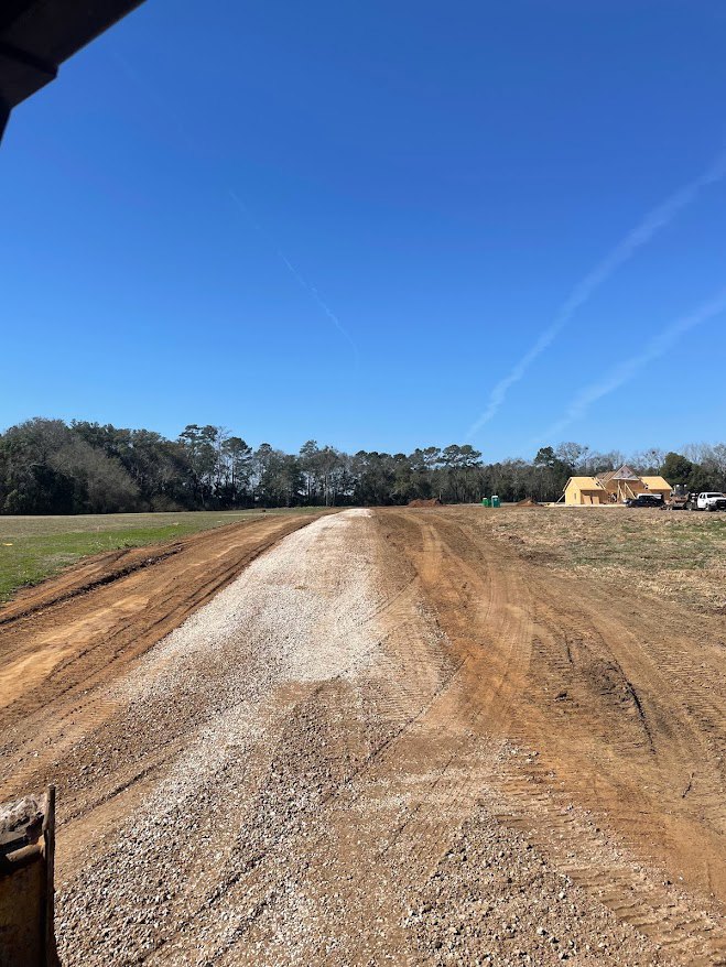 Dirt road bordered by tall trees leading to a house in the background, blue sky with scattered clouds and a jet trail overhead, grassy soil and wooden post visible in foreground