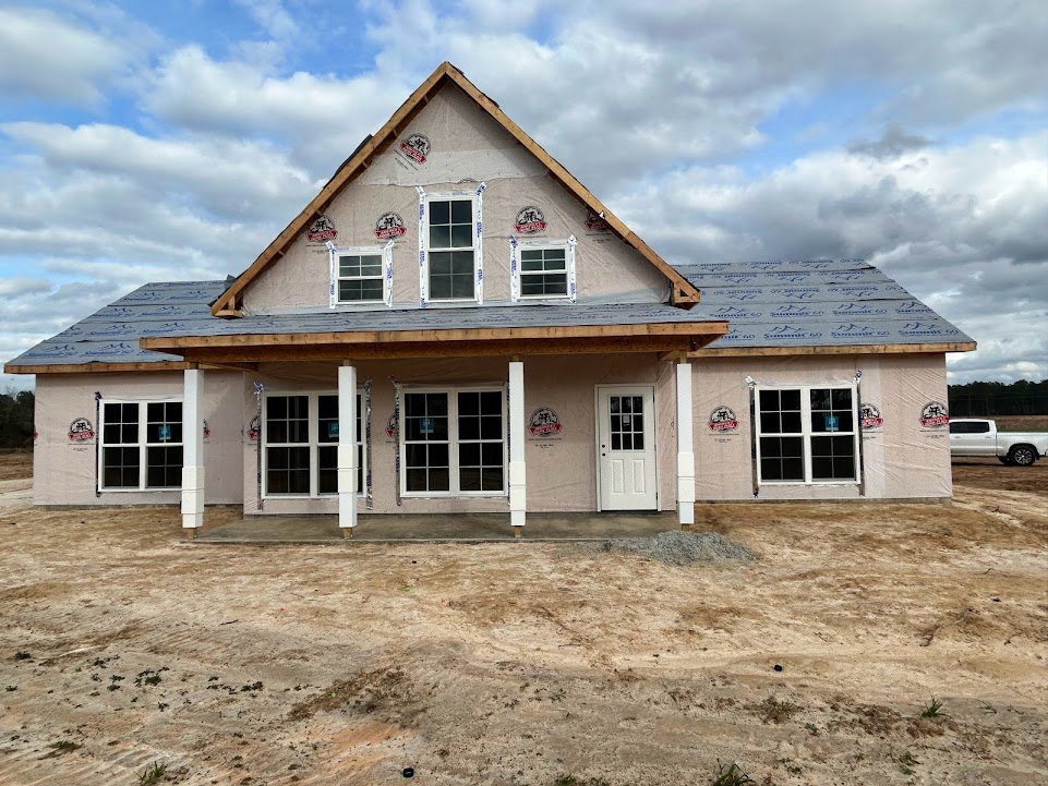 Two-story house under construction with white framed windows, multi-pane glass door, dirt patch in front yard, white truck parked on road, cloudy sky overhead