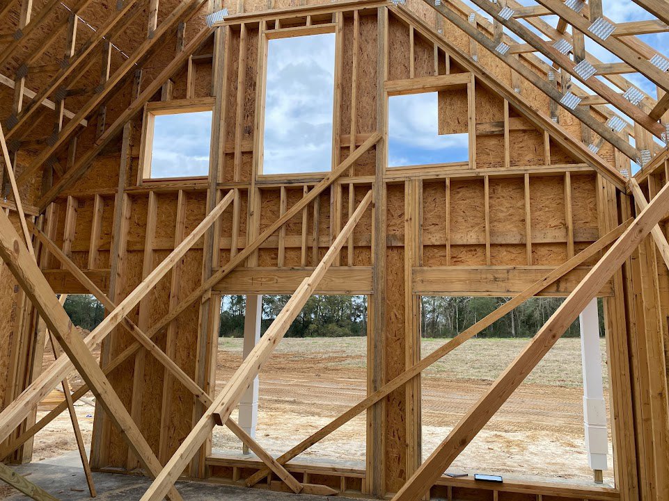 Exposed wooden beams and framing structure of a house under construction, open window cutouts, white support pillar with brown base, clear sky with clouds visible through window