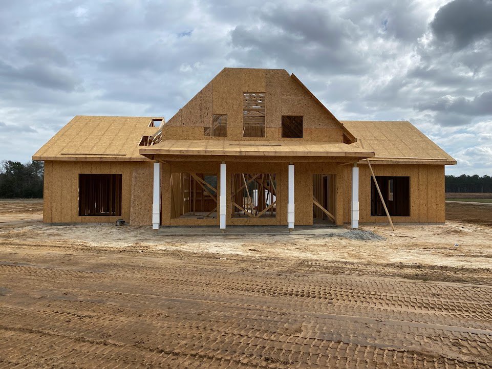 Framed house under construction with exposed wood, dirt lot in foreground, cloudy sky overhead