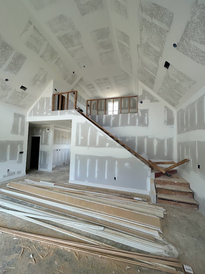 Open-concept room featuring unfinished wooden staircase, white painted ceiling, exposed wood planks, and plastered walls in a house under construction