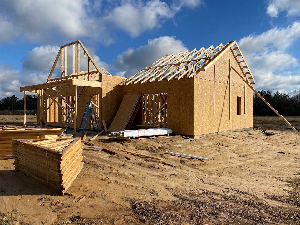 Wood-framed house under construction on a dirt lot with stacks of lumber, blue sky and scattered clouds overhead