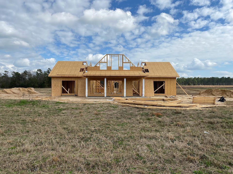 Wood-framed house under construction on grassy lot, exposed beams and plywood, blue sky with scattered clouds, trees in background