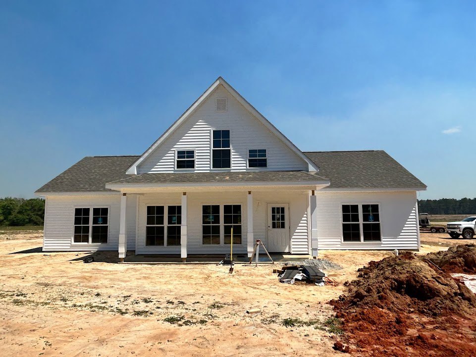 Partially built house with triangular roof, multi-pane window, exposed siding, ladder leaning against wall, white truck parked on dirt lot, blue sky overhead