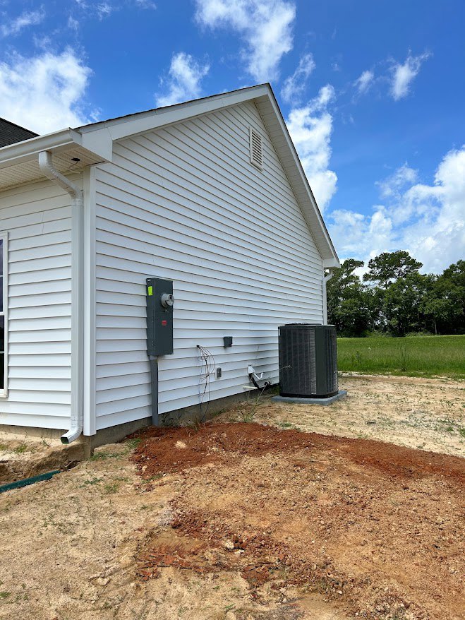 White house with gray box-shaped addition, black heat pump unit beside exterior wall, vent visible, surrounded by trees and grass, dirt patch in front yard.