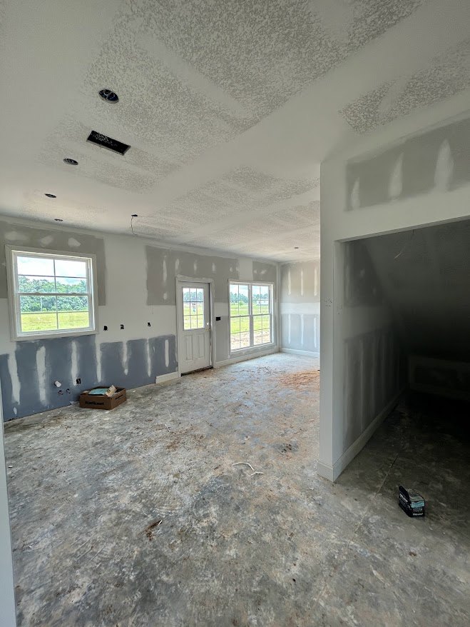 White-walled room with a white door featuring a window, large window overlooking grass and trees, plaster ceiling, dirty composite material floor.