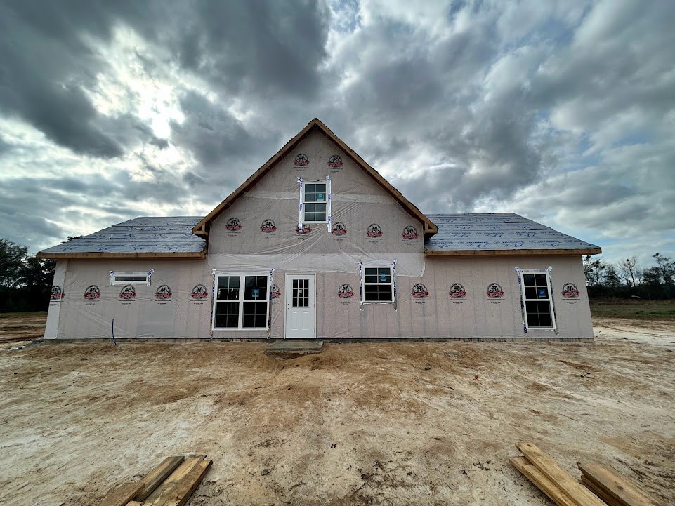 Partially built house with plastic sheeting covering exterior, white framed windows and glass paneled door, dirt ground in foreground, cloudy sky overhead
