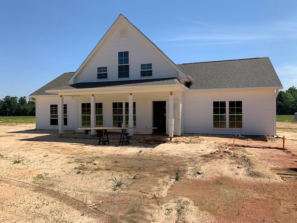 White custom home under construction with exposed dirt foreground, multi-pane windows, glass panel door, and blue sky background