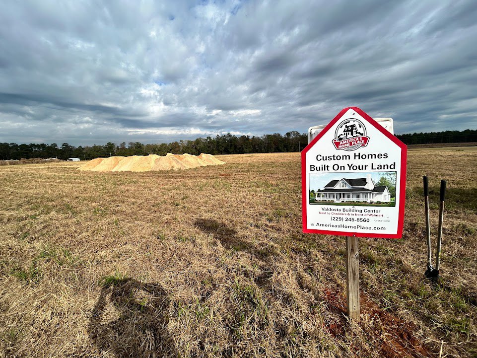 White house with black roof behind grassy field, wooden sign on post, cloudy sky, trees, and pile of sand in foreground