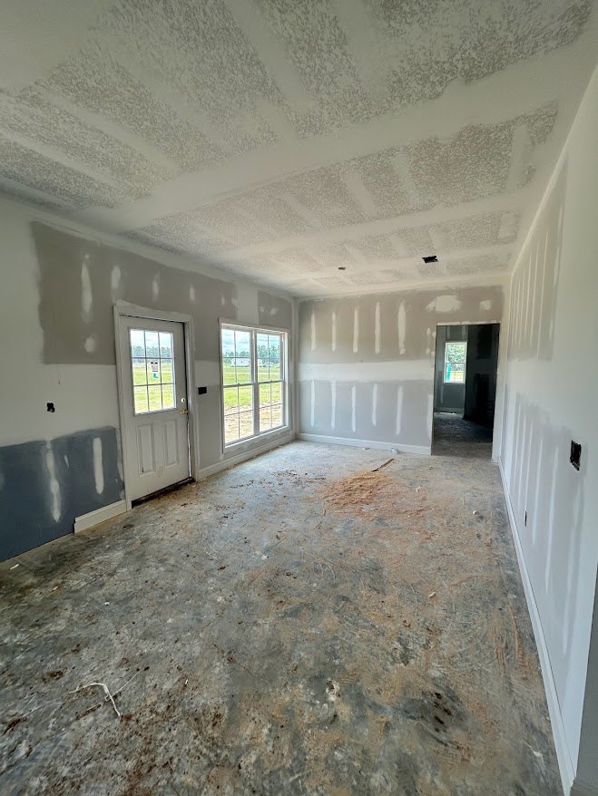 Sunlit room with white walls, large windows overlooking a grassy field, white baseboards, and a door; floor shows visible dirt and wear.