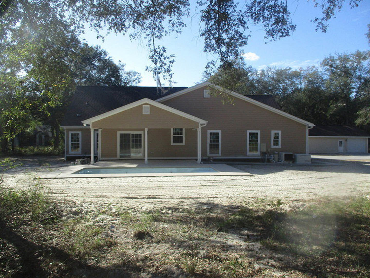 Modern home with white roof and wide overhang, sliding glass door and white-framed windows facing front yard, rectangular pool bordered by patio, tree branches partially shading