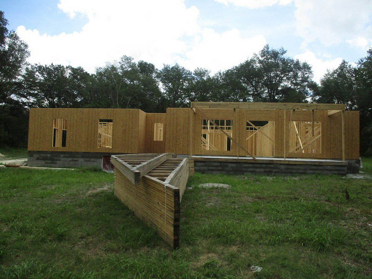 Wooden framing and beams of a house under construction, surrounded by grass and trees under a partly cloudy sky