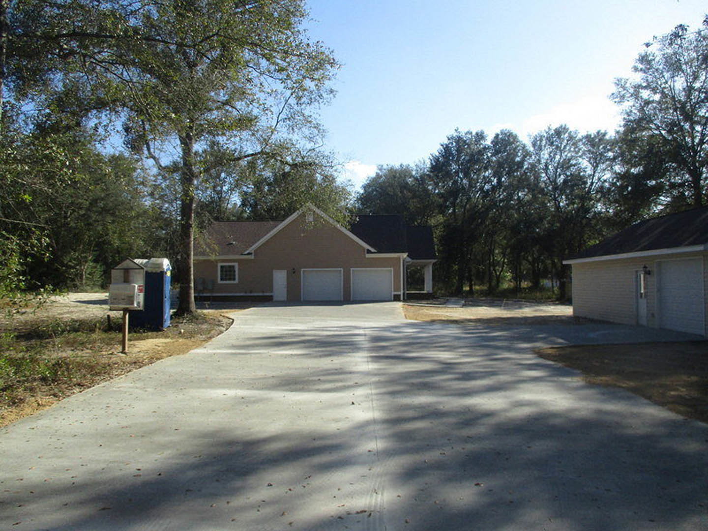Two-story home with attached garage, light-colored siding, paved driveway, mature trees in the background, manicured lawn, and clear sky overhead