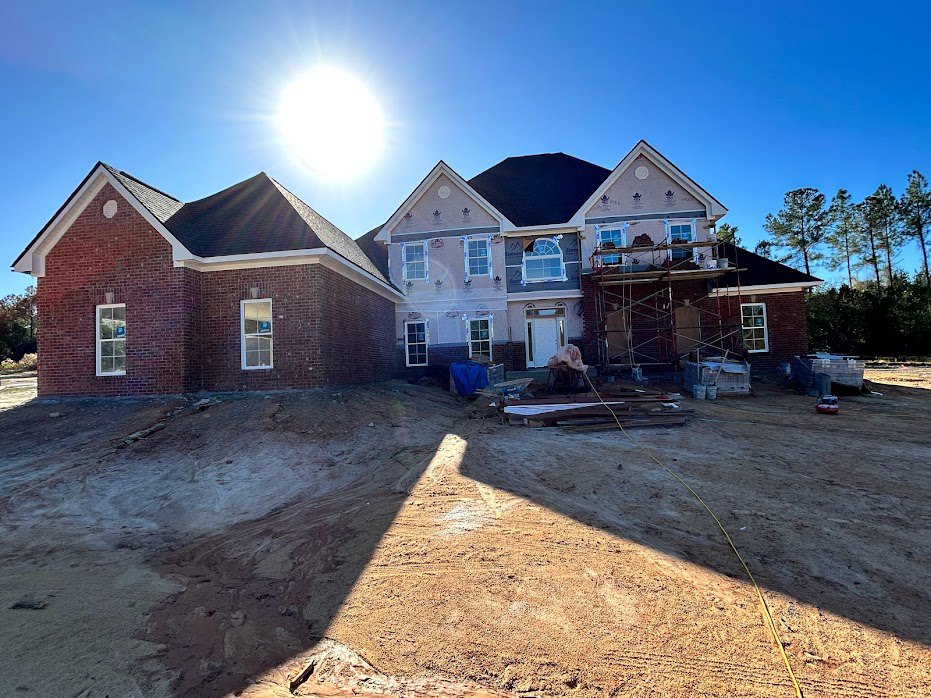 Partially built brick house with exposed framing, dirt construction site in foreground, clear blue sky overhead, scattered trees in background