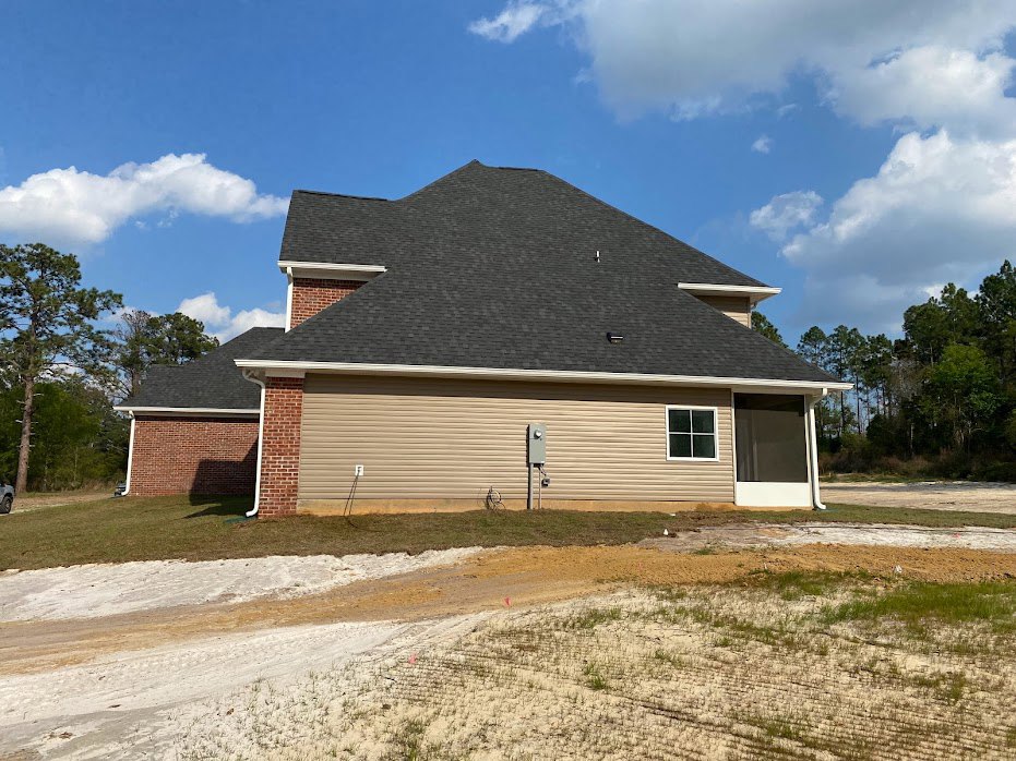 Two-story house with white siding, gray shingle roof, attached garage, screened window, white-framed windows, dirt road bordered by wooden fence, trees with green leaves in the