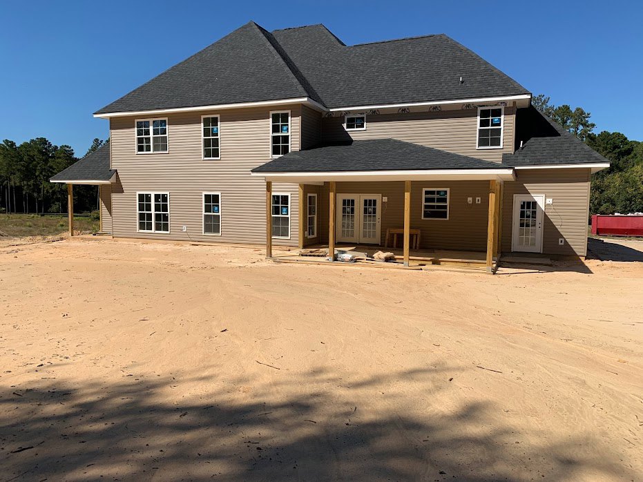 Framed custom home under construction with black roof, white trim, double glass-paneled doors, dirt foreground, and trees in the background under a blue sky