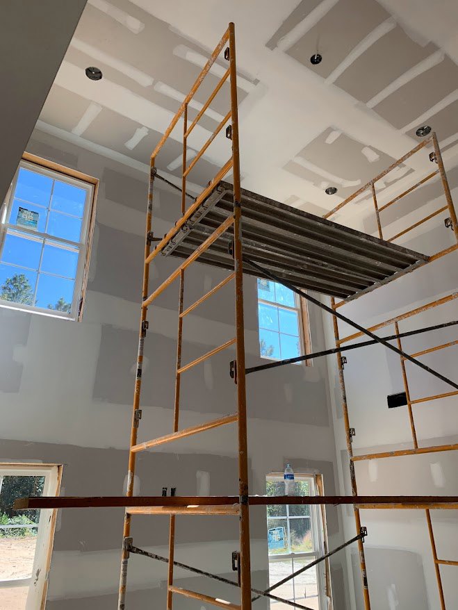 Scaffolding and wooden ladder set up in a partially finished room with exposed wood beam, large window showing blue sky, and construction sign taped to glass