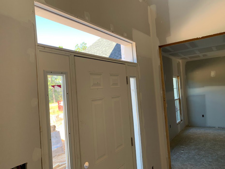 White door with glass window, concrete floor featuring a square border, large window showing roof and sky, white walls with ceiling molding, modern interior finishes.