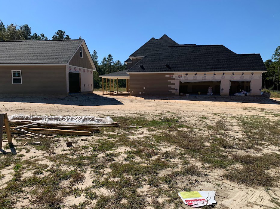 Wood-framed house under construction with exposed roof trusses, open garage, and construction materials scattered on grassy field