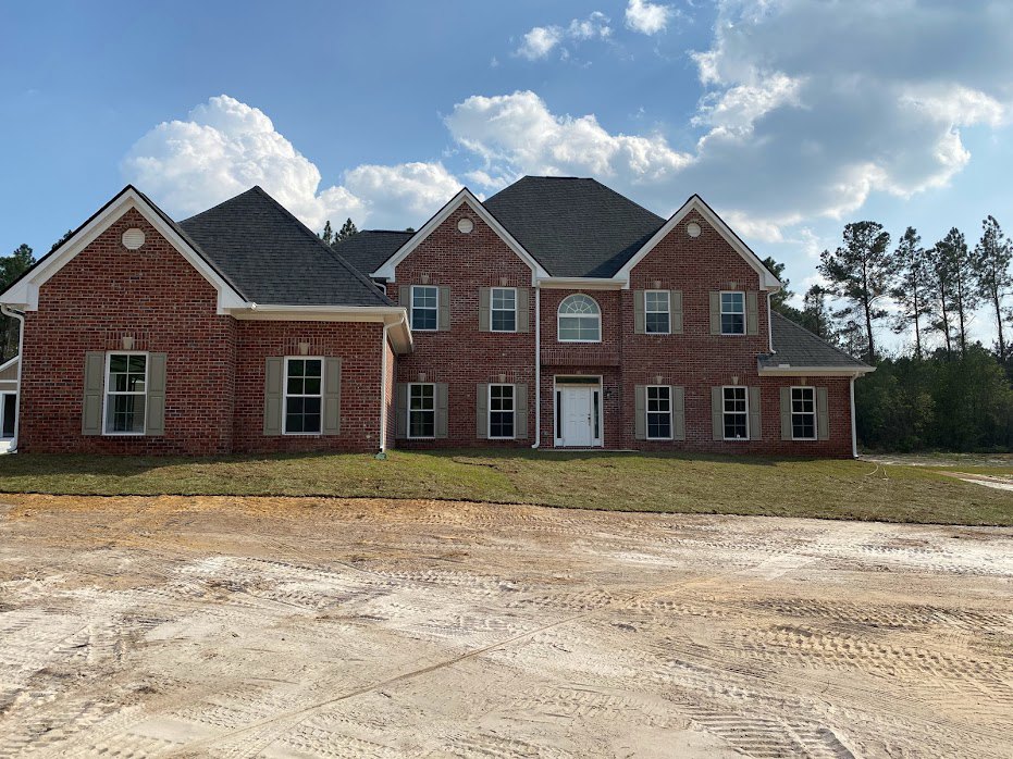 Red brick house with white door and glass panes, driveway with tire tracks, large window, trees in background, cloudy sky