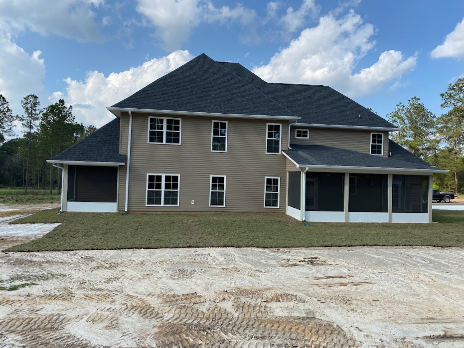 Two-story house with white-framed windows, covered porch, shingle roof, grass lawn, dirt driveway with tire tracks, blue sky and scattered clouds