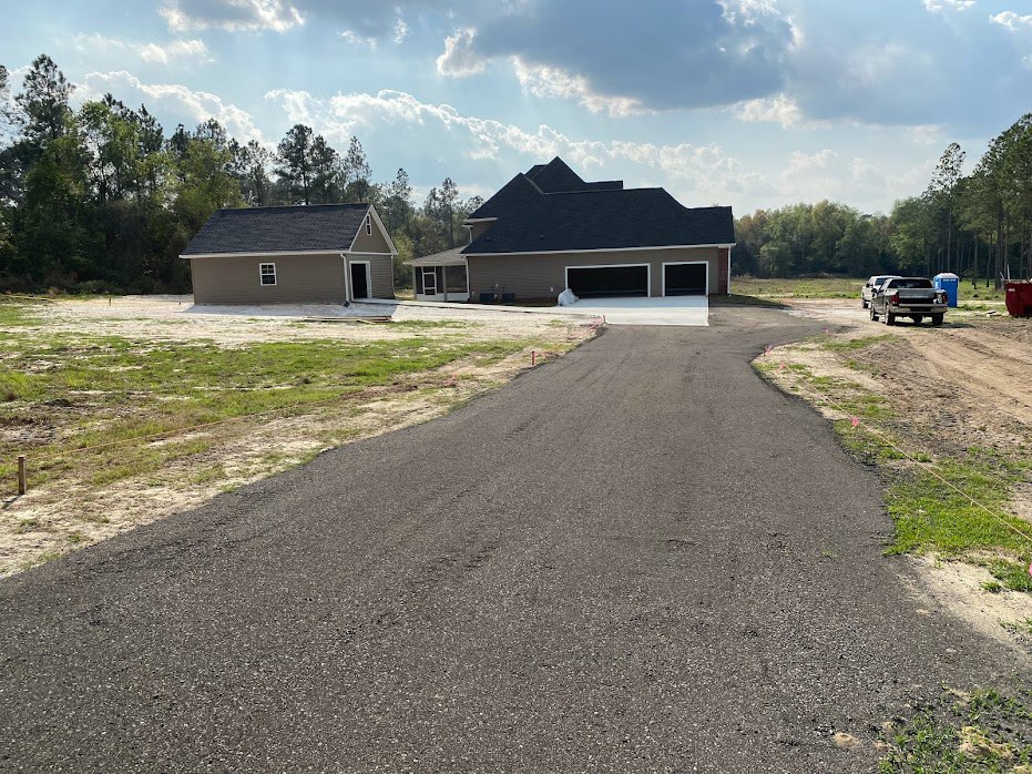 Paved driveway bordered by green grass and trees leading to a house with a white roof, attached garage, and blue sky with scattered clouds overhead